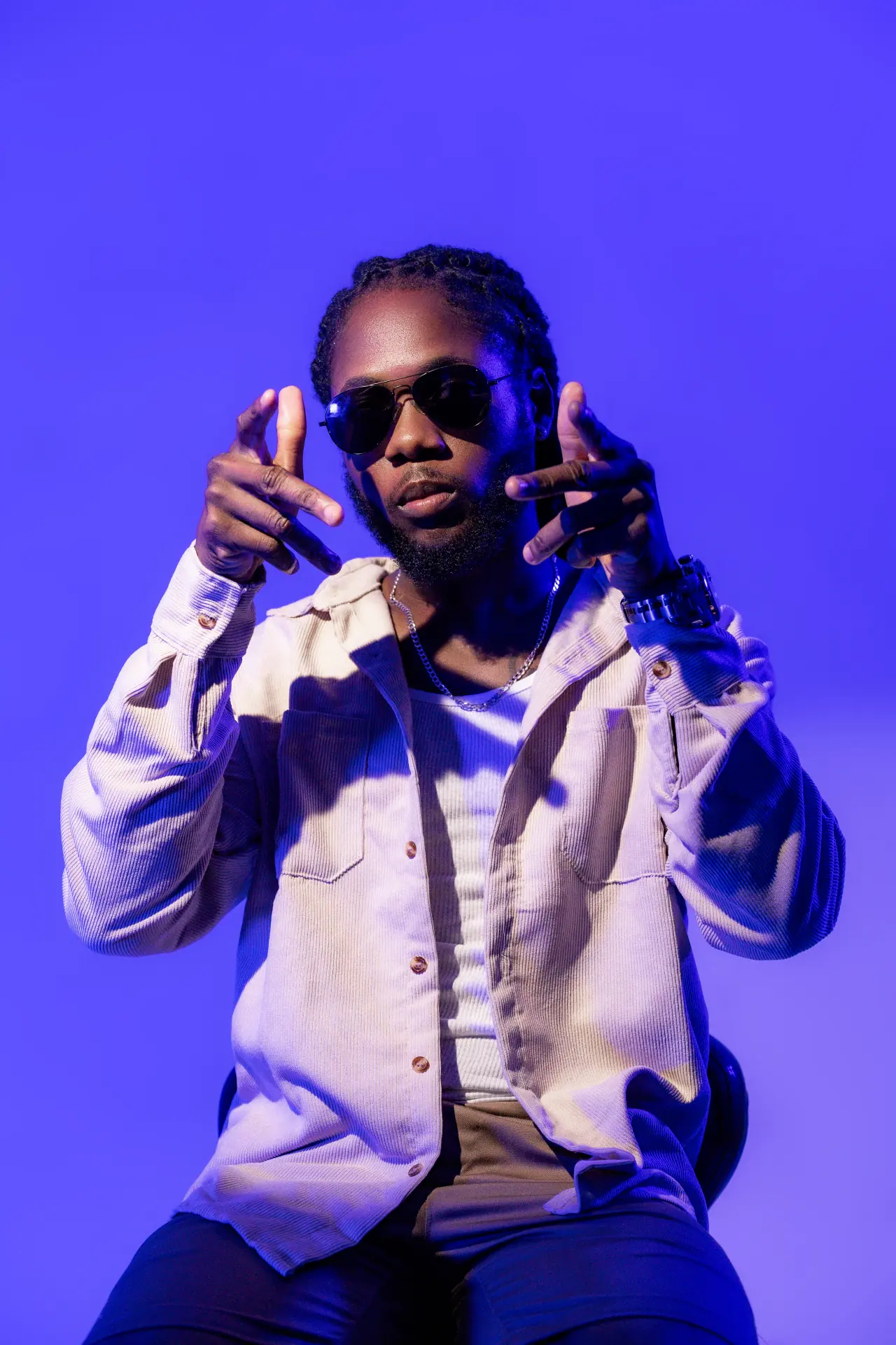 Saint Chris seated in blue light wearing sunglasses and a cream overshirt.